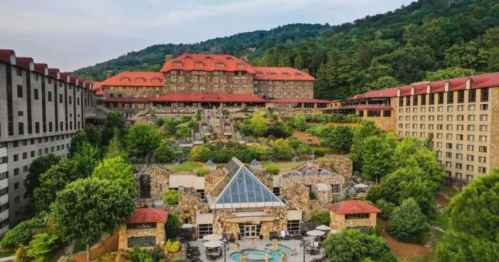 A scenic view of a large hotel complex surrounded by lush greenery and mountains, featuring a central pool area.