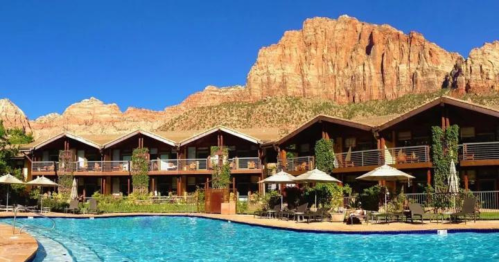 A resort with a pool in the foreground, surrounded by mountains and lush greenery under a clear blue sky.