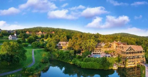 A scenic view of a resort by a lake, surrounded by lush greenery and blue skies with fluffy clouds.