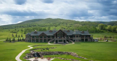 A large, luxurious lodge surrounded by green hills and trees, with a stone pathway and outdoor seating area in front.
