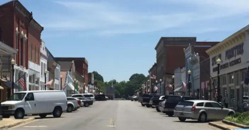 A quiet street lined with historic buildings, parked cars, and American flags under a clear blue sky.