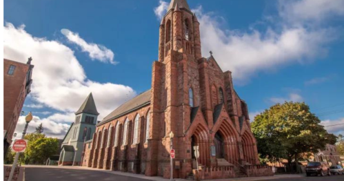 Historic stone church with tall spires, surrounded by trees and a blue sky with clouds.