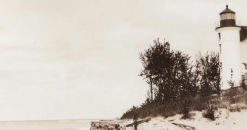 A vintage black-and-white photo of a lighthouse by the shore, surrounded by trees and sandy beach.