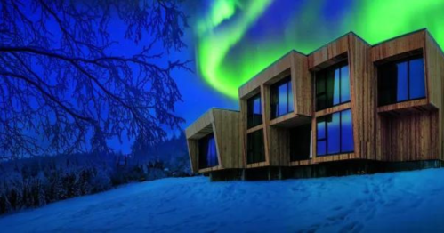 A modern wooden building under a vibrant aurora borealis in a snowy landscape at night.
