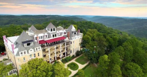 Aerial view of a grand mansion surrounded by lush greenery and rolling hills under a colorful sky.