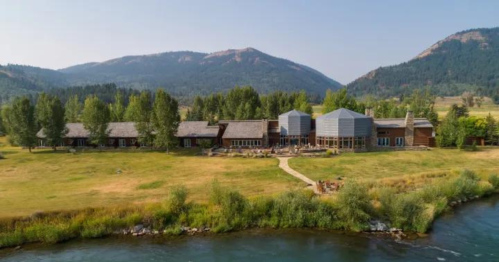 A scenic view of a lodge by a river, surrounded by trees and mountains under a clear blue sky.