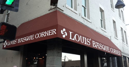 Sign for Louis' Basque Corner on a building with a maroon awning and white brick walls.