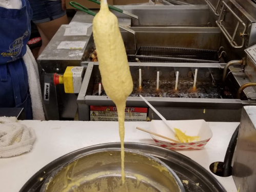A corn dog being dipped in batter, with a fryer in the background and a tray of condiments nearby.