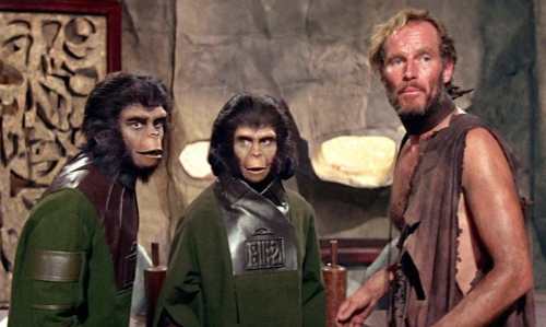 A human and two ape-like characters in a cave setting, wearing green uniforms, looking curiously at the viewer.