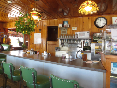 A cozy diner interior with wooden walls, a counter with green stools, and vintage decor, including lamps and a clock.