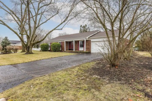 A single-story brick house with a driveway, surrounded by trees and a grassy lawn on a cloudy day.