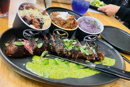 A plate of sliced steak with green sauce, accompanied by three bowls of sides: pasta, chili, and purple mashed potatoes.