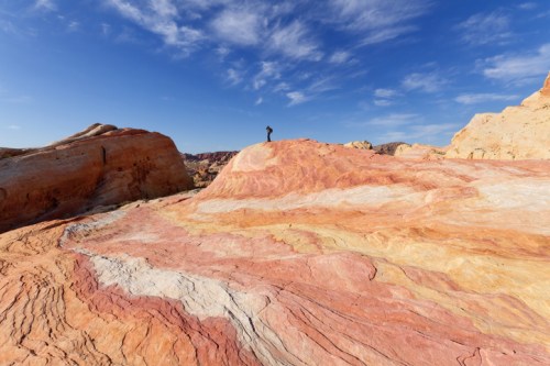 A person stands on colorful, layered rock formations under a blue sky with scattered clouds.