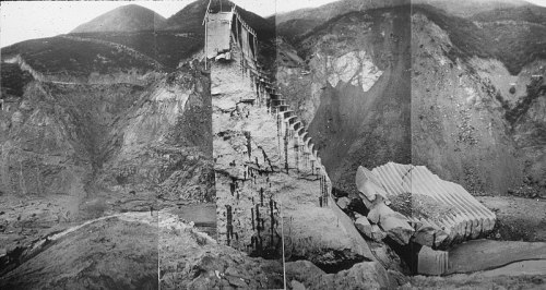 A black and white photo of a collapsed dam, with eroded land and debris scattered around the site.