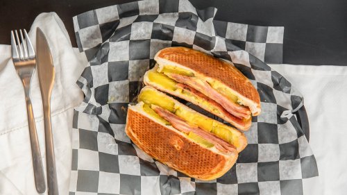 A sandwich with ham and pickles, cut in half, on a black and white checkered paper with utensils beside it.