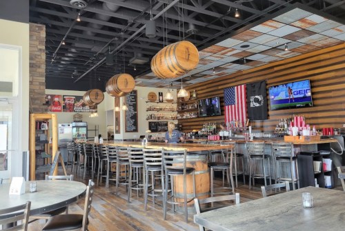 A modern bar interior featuring wooden barrels, a long counter, and a TV, with rustic decor and a warm atmosphere.