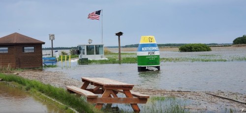 A flooded area with a picnic table, a buoy marked "Northern Point," and an American flag in the background.