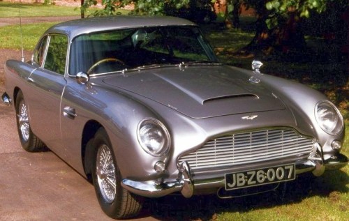 A classic silver Aston Martin sports car parked on a driveway, surrounded by greenery.