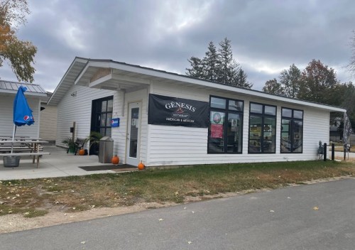 A white building with large windows, featuring a sign for "Genesis" and pumpkins outside, set against a cloudy sky.