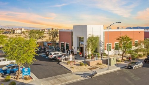 Aerial view of a modern building with a line of people outside, surrounded by desert landscape and parked cars.