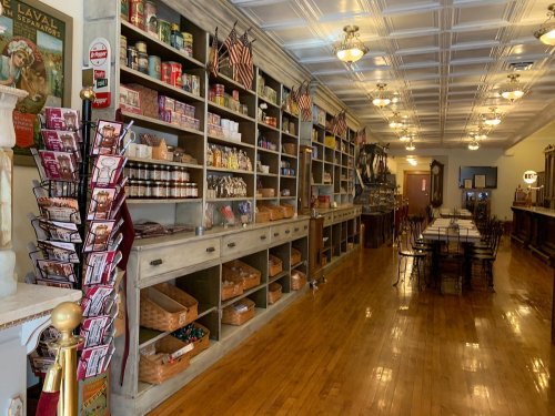 A vintage store interior with wooden shelves filled with jars and cans, and a long dining table in the center.