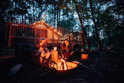 A cozy campfire scene with two people sitting by the fire, surrounded by trees and a cabin in the background.