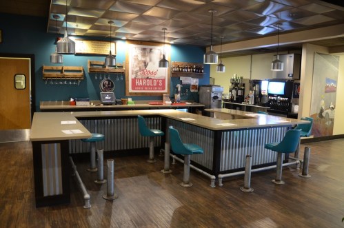 A retro diner interior featuring a counter with teal stools, metal accents, and vintage decor on the walls.