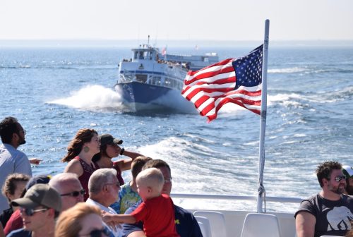 A boat sails on the water, with passengers enjoying the ride and an American flag waving in the foreground.