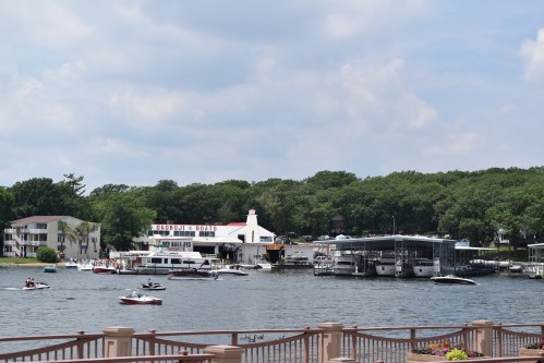 A scenic view of a marina with boats, a waterfront building, and lush greenery under a partly cloudy sky.