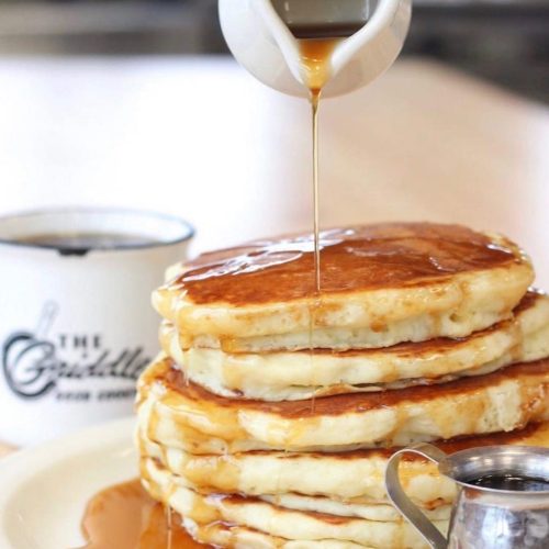 A stack of fluffy pancakes drizzled with syrup, with a cup in the background.