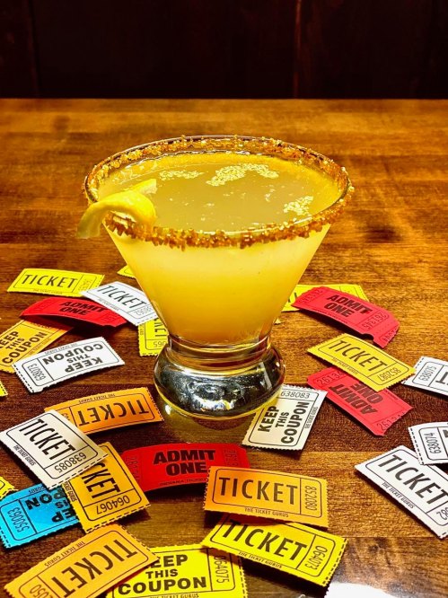 A cocktail with a lemon slice sits on a wooden table surrounded by colorful ticket stubs.