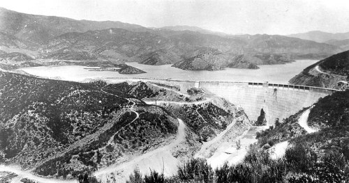 Historic black-and-white photo of a dam surrounded by mountains and a lake, with winding roads in the foreground.