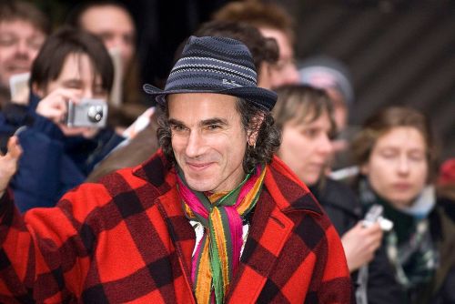 A man in a red and black checkered coat and hat waves to a crowd, smiling amidst onlookers with cameras.