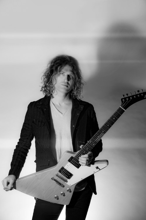 A musician with curly hair stands confidently holding a white electric guitar, dressed in a black jacket against a soft backdrop.