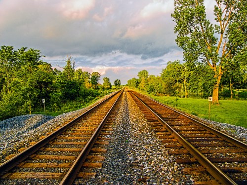 A view of parallel railway tracks stretching into the distance, surrounded by lush greenery and a cloudy sky.