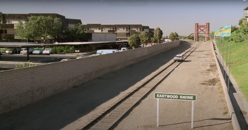 A view of Eastwood Ravine with a road, a sign, and buildings in the background under a clear sky.