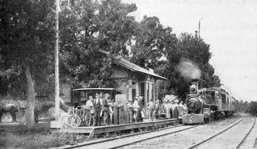A vintage train at a station with passengers, bicycles, and a building surrounded by trees.