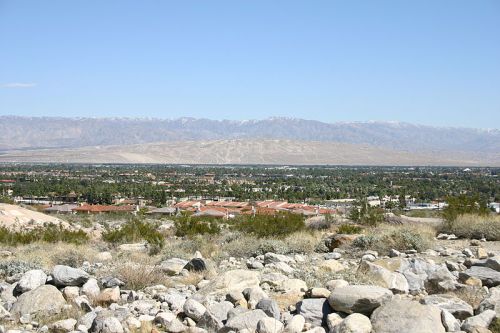 A panoramic view of a valley with rocky terrain, greenery, and distant mountains under a clear blue sky.