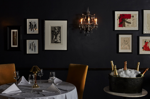 Elegant dining setup with a round table, wine glasses, and a bucket of champagne, against a dark wall with framed art.