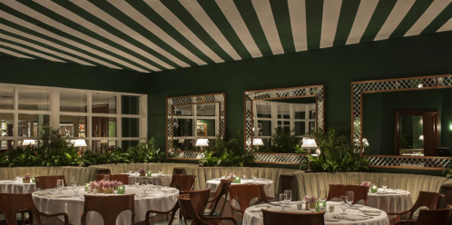 Elegant restaurant interior with green walls, striped ceiling, round tables, and lush plants. Mirrors reflect the ambiance.