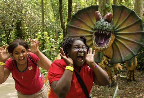 Two excited women pose playfully in a lush forest, with a colorful dinosaur statue behind them.