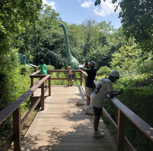 A wooden walkway in a park with people observing dinosaur sculptures surrounded by greenery.