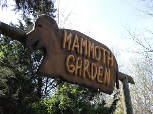Wooden sign for "Mammoth Garden" featuring a mammoth silhouette, surrounded by trees and greenery.