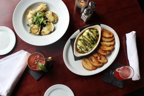 A table setting with two dishes: one with stuffed pasta and salad, the other with baked cheese and toasted bread, plus drinks.