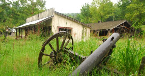 An old rustic wagon wheel and pipe in overgrown grass near abandoned wooden buildings at Dogpatch USA in Arkansas.
