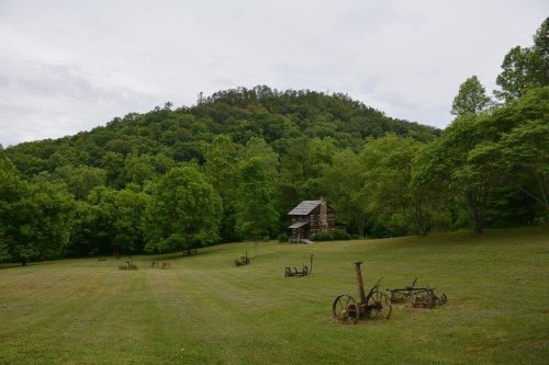 A rustic cabin surrounded by lush green trees and a grassy field, with old farming equipment scattered in the foreground.
