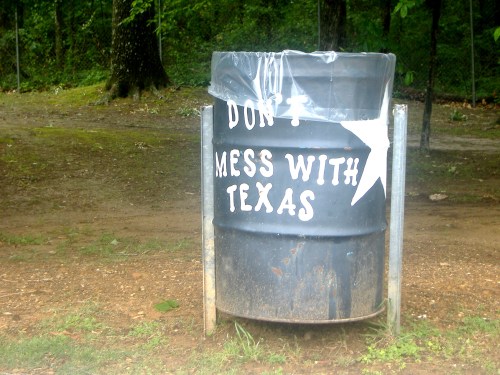 A trash can with a sign that reads "Don't Mess With Texas," set in a wooded area.