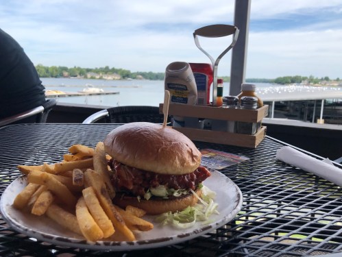 A plate with a burger and fries sits on a table by a lake, with boats in the background and condiments nearby.