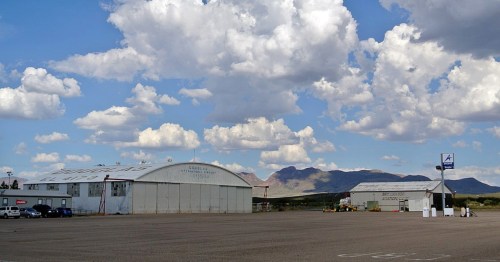 A small airport with hangars under a blue sky filled with clouds, surrounded by mountains in the background.