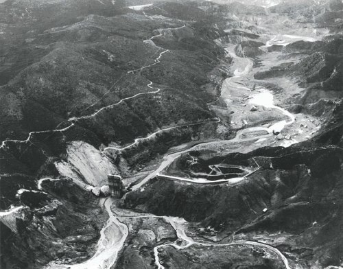 Aerial view of a mountainous landscape with a dam and a riverbed, showcasing erosion and construction activity.